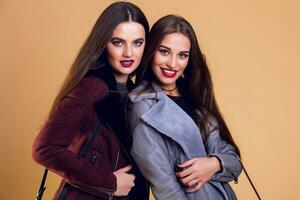 Two sexy models in casual trendy spring outfit staying back to back indoor in studio. Long brunette hairstyle , high fashion look, bright red lips. photo