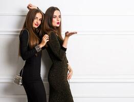 two elegant young women in glamour bleach long dress posing on white wall , enjoying event , send kiss,  trendy hand bag , red lips. New year or birthday party. photo