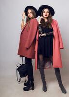 Full height   studio image of two fashionable sexy woman in casual trendy spring coat,  boots with heels , black hat, stylish hand bag. photo