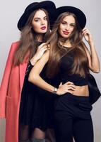 Fashion image of two girls, sisters, posing on grey background, hugging, smiling. Wearing stylish pink  coat and black hat, short top and evening dress. photo