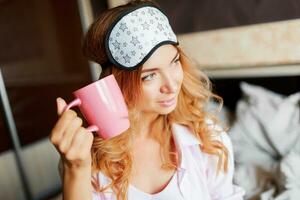 Pretty woman with candid smile posing in eye mask  at home and drinking hot tea. Cozy home atmosphere. photo