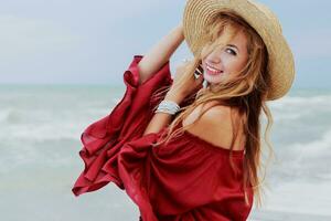 Close up portrait of smiling pretty woman in straw hat and trendy red summer dress posing near ocean waves. Windy  and cloudy weather. photo