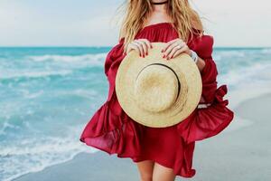White  ginger  woman in stylish dress  holding straw hat and  posing  on the beach near ocean. Blu sky. Windy weather photo