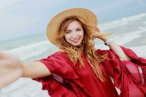 Happy smiling  white ginger  woman in stylish  red dress  making self portrait mobile phone  on the beach near ocean. Wearing straw hat.  Shows signs by hand. photo