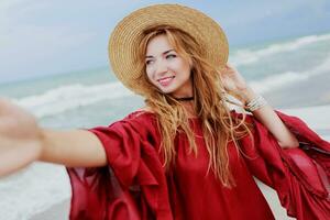 Happy smiling  white ginger  woman in stylish  red dress  making self portrait mobile phone  on the beach near ocean. Wearing straw hat.  Shows signs by hand. photo