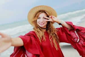 Happy smiling  white ginger  woman in stylish  red dress  making self portrait mobile phone  on the beach near ocean. Wearing straw hat.  Shows signs by hand. photo