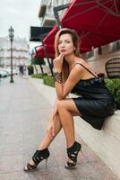 Slim tanned woman in elegant black dress and heels with bright blonde hairs posing in old European city near luxury restaurant. photo