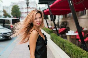 Positive woman in black dress walking on streets, enjoying vacation. Windy hairs. photo