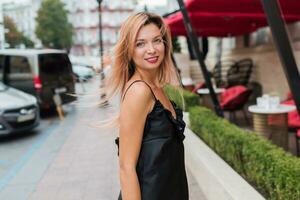 Positive woman in black dress walking on streets, enjoying vacation. Windy hairs. photo