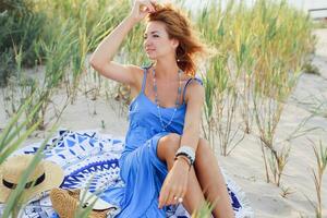 Shy  girl with perfect tan skin  posing on sunny beach in trendy blue dress, sitting on sand. Windy hairs. Evening sunlight. photo