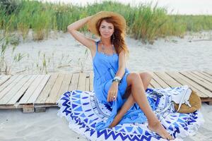 Smiling   redhead woman sitting on  beach towel .Perfect tan body. Blue dress. Windy hairs. photo