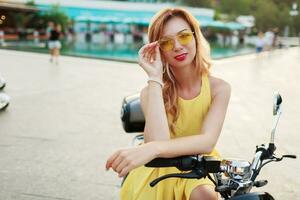 Cheerful ginger woman in yellow glasses and vintage dress sitting on stylish motorbike. Vacation mood. Sunny summer day. Red lips. photo