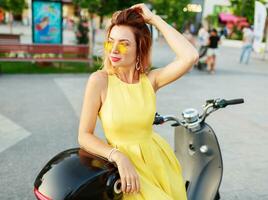 Smiling   ginger  woman in yellow dress riding by motorbike , traveling and having fun. Wearing stylish summer outfit. photo