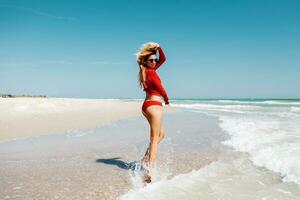 de viaje rubio mujer saltando con contento expresión , vueltas alrededor en agua. Oceano olas antecedentes. vistiendo rojo bikini. lleno longitud. verano vacaciones. foto