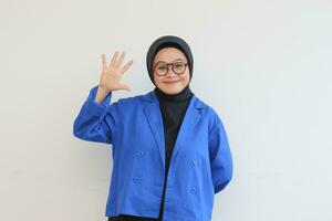 Beautiful young Asian Muslim woman, wearing glasses and blue blazer showing palms while smiling photo