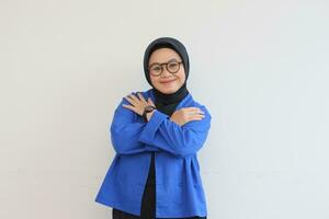 Young beautiful Asian Muslim woman, wearing glasses and blue blazer smiling hugging herself photo