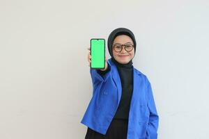 smiling beautiful asian women in blue blazer and hijab showing green screen mobile phone photo