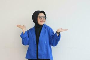 Beautiful young Asian Muslim woman in glasses, hijab and wearing blue blazer smiling confident and happy with both hands pointing up photo