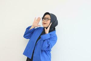 beautiful young Asian Muslim woman, wearing glasses and blue blazer with awe gestures while smiling photo