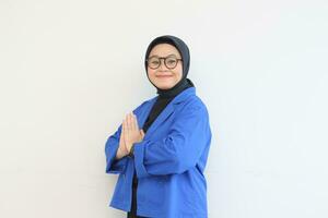 beautiful young Asian Muslim woman, wearing glasses and blue blazer with namaste gesture while smiling photo