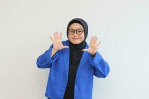 Beautiful young Asian Muslim woman, wearing glasses and blue blazer showing palms while smiling photo