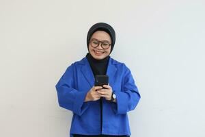 beautiful young asian woman in glasses, hijab and wearing blue blazer looking at her mobile phone with happy face photo