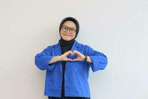 hermosa joven asiático mujer vistiendo anteojos, hijab y vistiendo azul chaqueta de sport gesticulando amor firmar con su mano foto