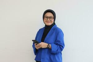 Beautiful young Asian woman in glasses, hijab and wearing a blue blazer is holding a mobile phone with a smiling face photo