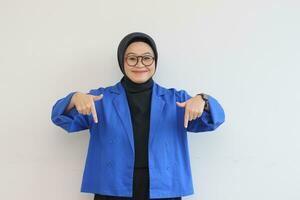 Beautiful young Asian Muslim woman in glasses, hijab and wearing blue blazer smiling confident and happy with both hands pointing down photo
