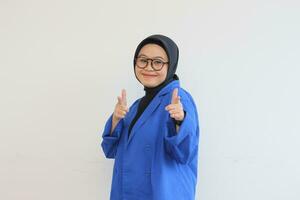 Beautiful young Asian Muslim woman in glasses, hijab and wearing blue blazer smiling confident and excited with both hands pointing forward photo