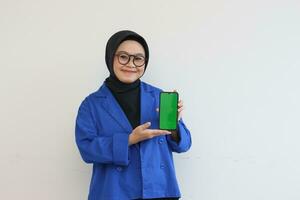 beautiful young asian woman in glasses, hijab and wearing blue blazer showing green screen mobile phone while smiling photo