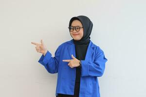 Beautiful young Asian Muslim woman in glasses, hijab and wearing blue blazer smiling confident and happy with both hands pointing sideways photo