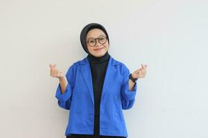 beautiful young asian woman wearing glasses, hijab and wearing blue blazer gesturing love sign with her hand photo