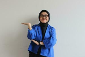 hermosa joven asiático musulmán mujer, vistiendo lentes y azul chaqueta de sport demostración palmas mientras sonriente foto
