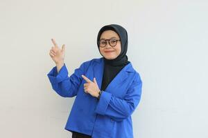 Beautiful young Asian Muslim woman in glasses, hijab and wearing blue blazer smiling confident and happy with both hands pointing up photo