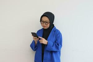 beautiful young asian woman in glasses, hijab and wearing a blue blazer looking at her mobile phone photo