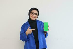 beautiful young asian woman in glasses, hijab and wearing blue blazer is holding green screen mobile phone with happy face photo