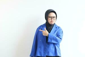 Beautiful young Asian Muslim woman in glasses, hijab and wearing blue blazer angry expression with hand pointing sideways photo