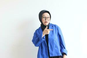 Beautiful young Asian Muslim woman in glasses, hijab and wearing blue blazer smiling confident and happy with both hands pointing sideways photo