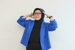 Beautiful young Asian Muslim woman, wearing glasses and blue blazer showing stress gesture while holding head photo