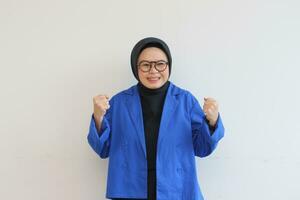 beautiful young Asian Muslim woman, wearing glasses and blue blazer with happy expression while clenching fist photo