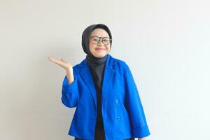Beautiful young Asian Muslim woman, wearing glasses and blue blazer showing palms while smiling photo