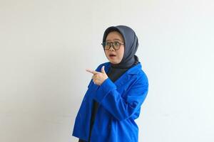 Beautiful young Asian Muslim woman in glasses, hijab and wearing blue blazer smiling confident and happy with both hands pointing sideways photo