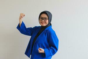 hermosa joven asiático musulmán mujer, vistiendo lentes y azul chaqueta de sport con contento expresión mientras apretando puño foto
