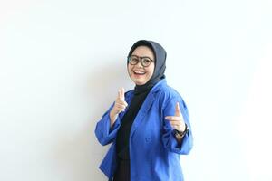 Beautiful young Asian Muslim woman in glasses, hijab and wearing blue blazer smiling confident and excited with both hands pointing forward photo