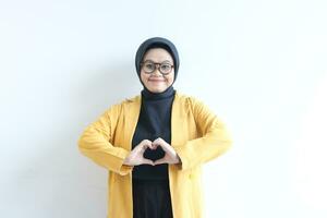 beautiful young asian woman wearing glasses, hijab and wearing yellow blazer gesturing love sign with her hand photo