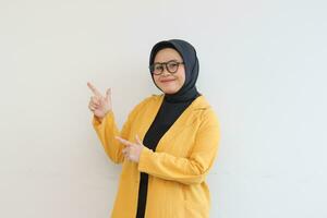 Beautiful young Asian Muslim woman in glasses, hijab and wearing yellow blazer smiling confident and happy with both hands pointing sideways photo