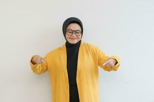 Beautiful young Asian Muslim woman in glasses, hijab and wearing yellow blazer smiling confident and happy with both hands pointing down photo