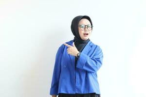 Beautiful young Asian Muslim woman in glasses, hijab and wearing blue blazer smiling confident and happy with hand pointing sideways isolated on white background, advertising concept photo