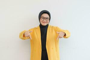 Beautiful young Asian Muslim woman in glasses, hijab and wearing yellow blazer smiling confident and happy with both hands pointing down photo